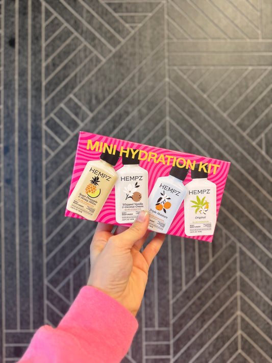 A hand wearing a pink sleeve holds a HEMPZ Mini Hydration Kit containing three small bottles against a gray geometric tiled wall.