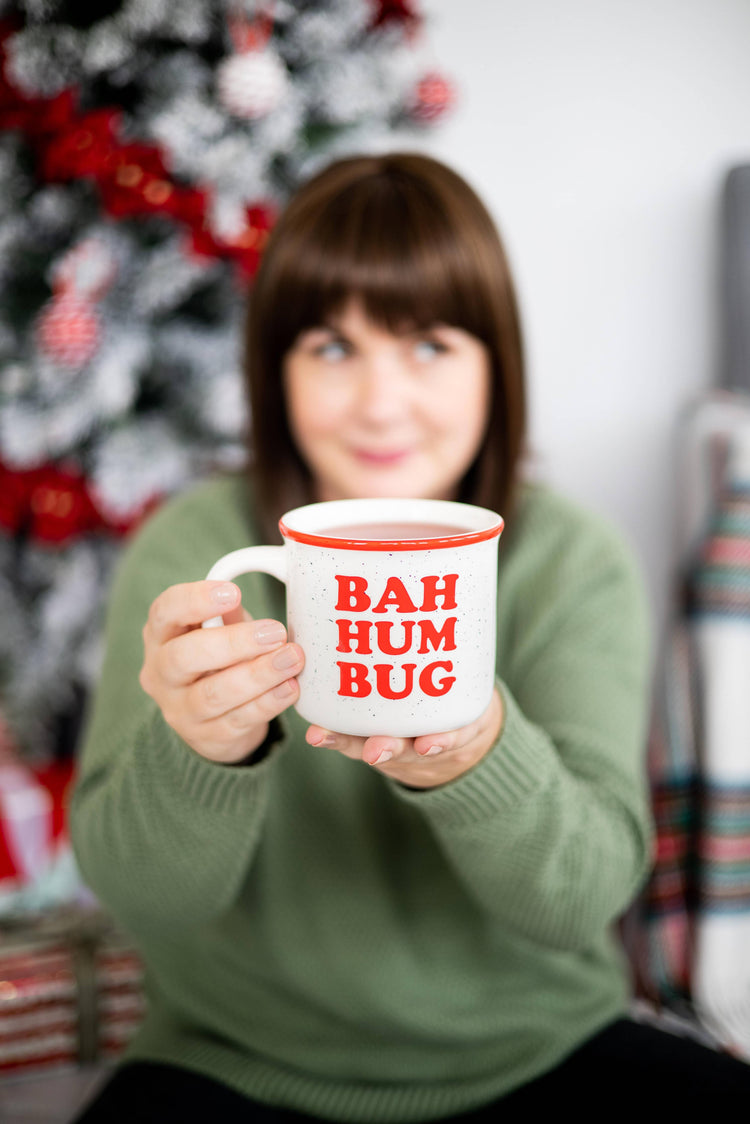 A woman in a green sweater holds up a white ceramic mug with red text reading 'BAH HUM BUG' in front of a blurred Christmas tree background.
