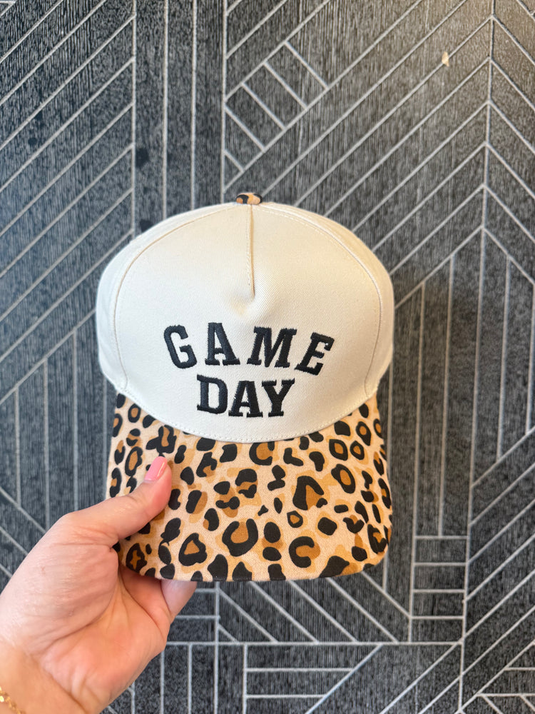 A hand holding a baseball cap with a cream-colored crown and a leopard-print visor. The front of the crown features the text 'GAME DAY' in black lettering.