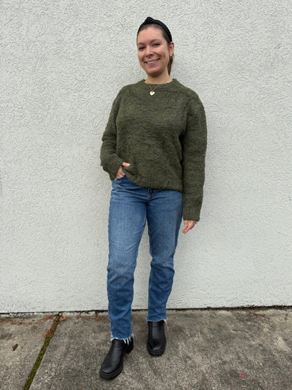 Person wearing a green sweater and blue jeans standing against a white wall.