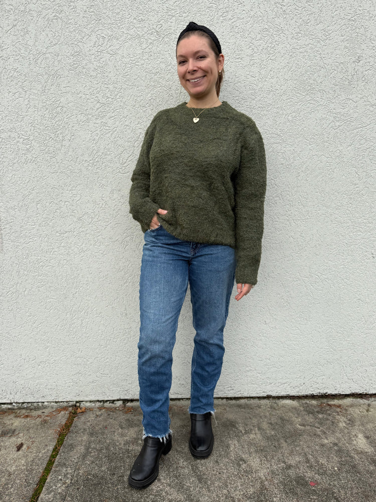 Person wearing a green sweater and blue jeans standing against a white wall.
