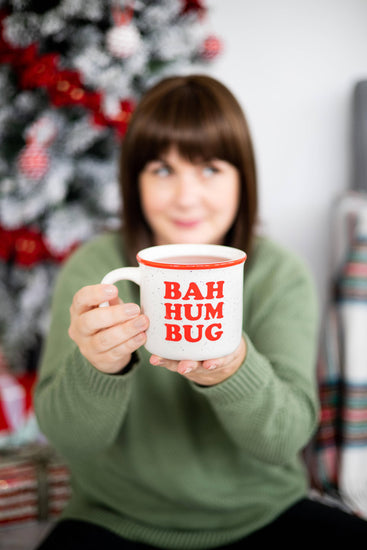 A woman in a green sweater holds up a white ceramic mug with red text reading 'BAH HUM BUG' in front of a blurred Christmas tree background.